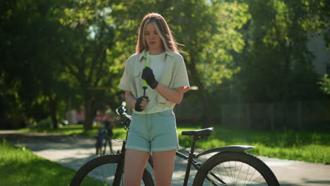 young girl with blonde hair stands in sunlight, focused while holding green air pump next to bicycle, background shows blurred figure walking and lush trees