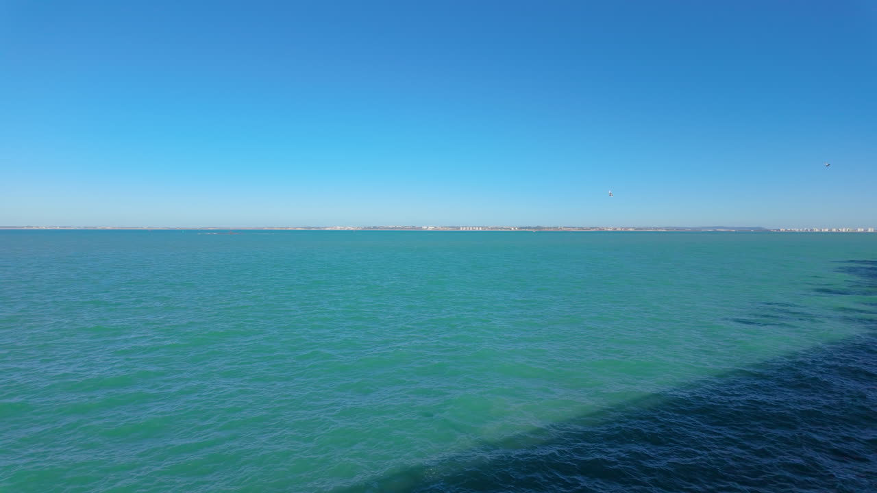 The expansive Bay of C&aacute;diz viewed from a seaside walkway, featuring tranquil turquoise waters meeting the clear blue sky at the horizon