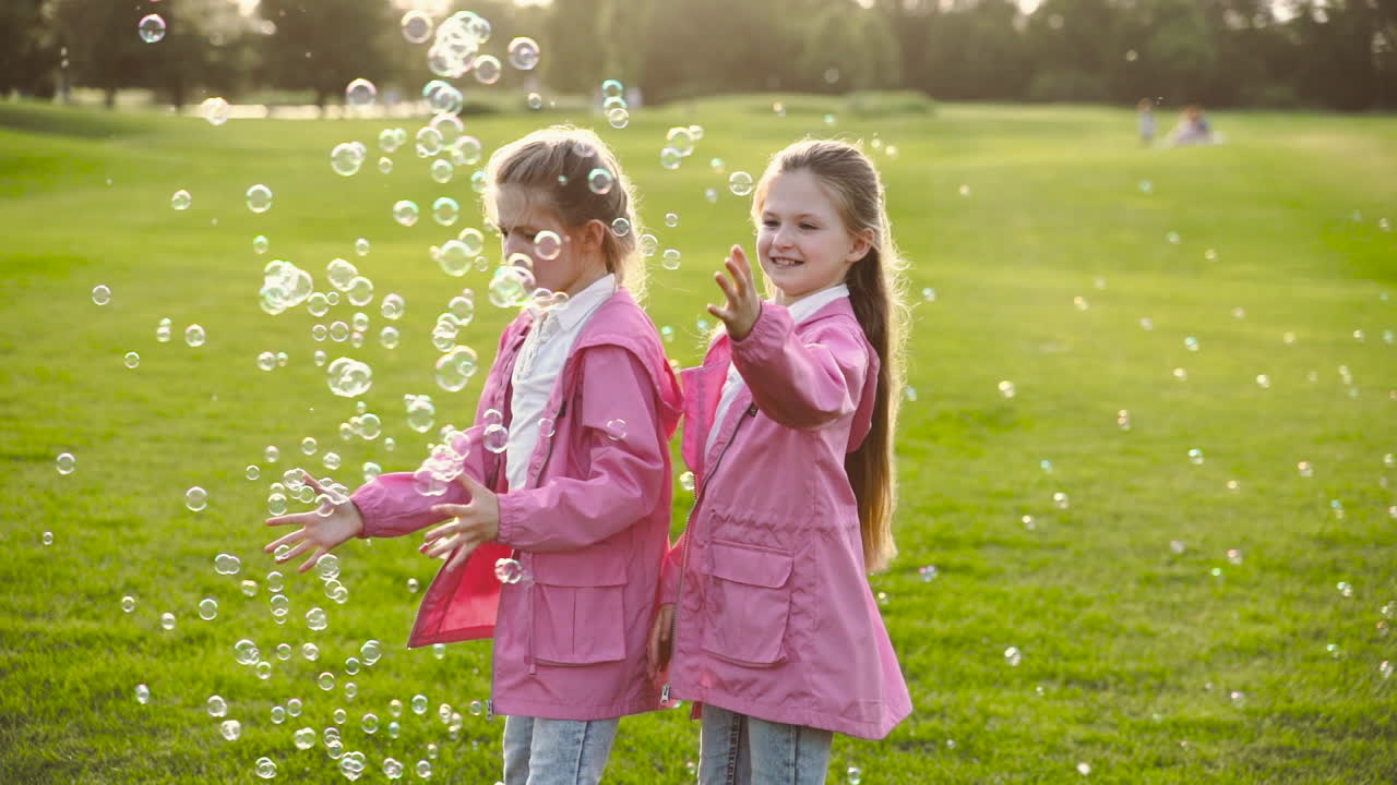똑같은 옷을 입은 행복한 작은 자매들이 비누방울을 잡고 손을 잡고 공원에서 빙글빙글 돌고 있다
