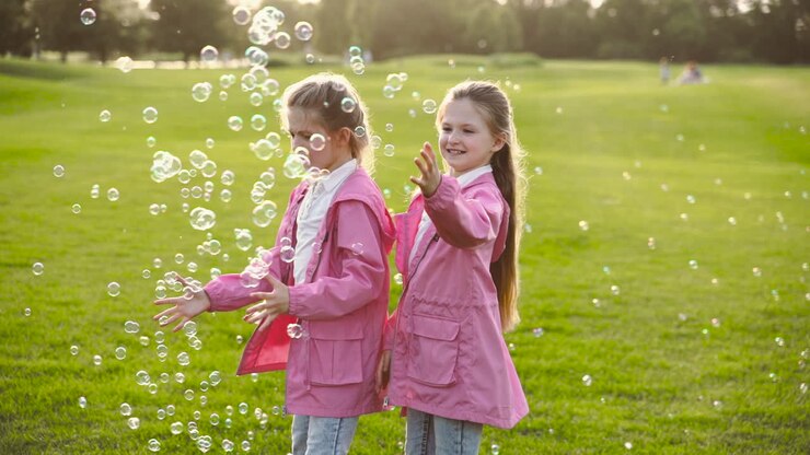 Sorelline felici con vestiti identici che acchiappano bolle di sapone, tenendosi per mano e volteggiando nel parco