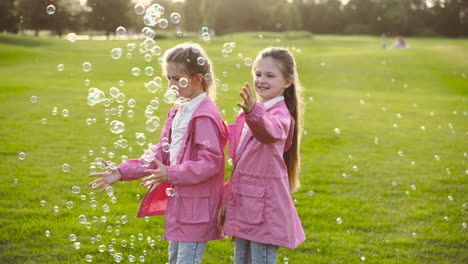 happy little sisters in identical clothes catching soap bubbles, holding hands and spinning around in the park