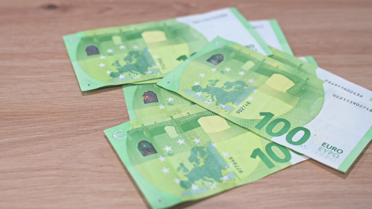 A close-up video of several €100 Euro banknotes spread across a wooden table, with a hand holding one of the bills in the foreground. The image emphasizes cash, wealth, and financial concepts.