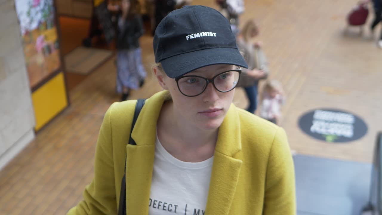 Young woman with feminist sign cap and glasses in the shopping mall going up with escalator and looking around