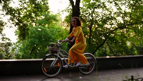 beautiful smiling woman in long yellow dress riding a city bicycle with a basket and flowers inside during the dawn. lens flare