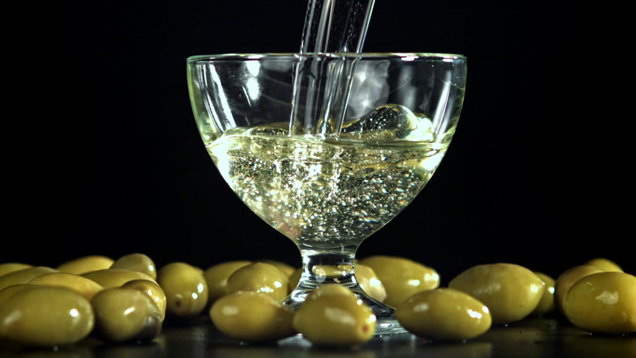 Versamento al rallentatore di liquido trasparente in un bicchiere con olive verdi