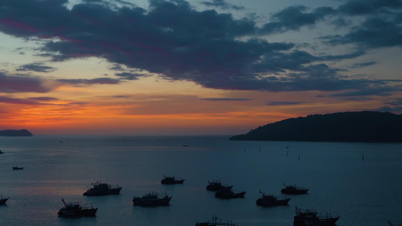 Aerial pan captures the serene Kota Kinabalu waterfront harbor with silhouetted fishing boats and islands against a stunning orange and blue sunset sky