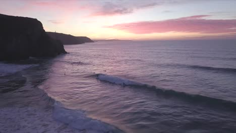 a cliff sits silhouetted as the sun sets and surfs play in the ocean in cornwall