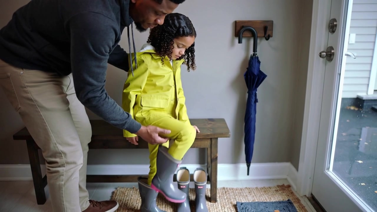 Seeing wet pavement dad helping child in yellow coat pulling on gray boots at entry bench
