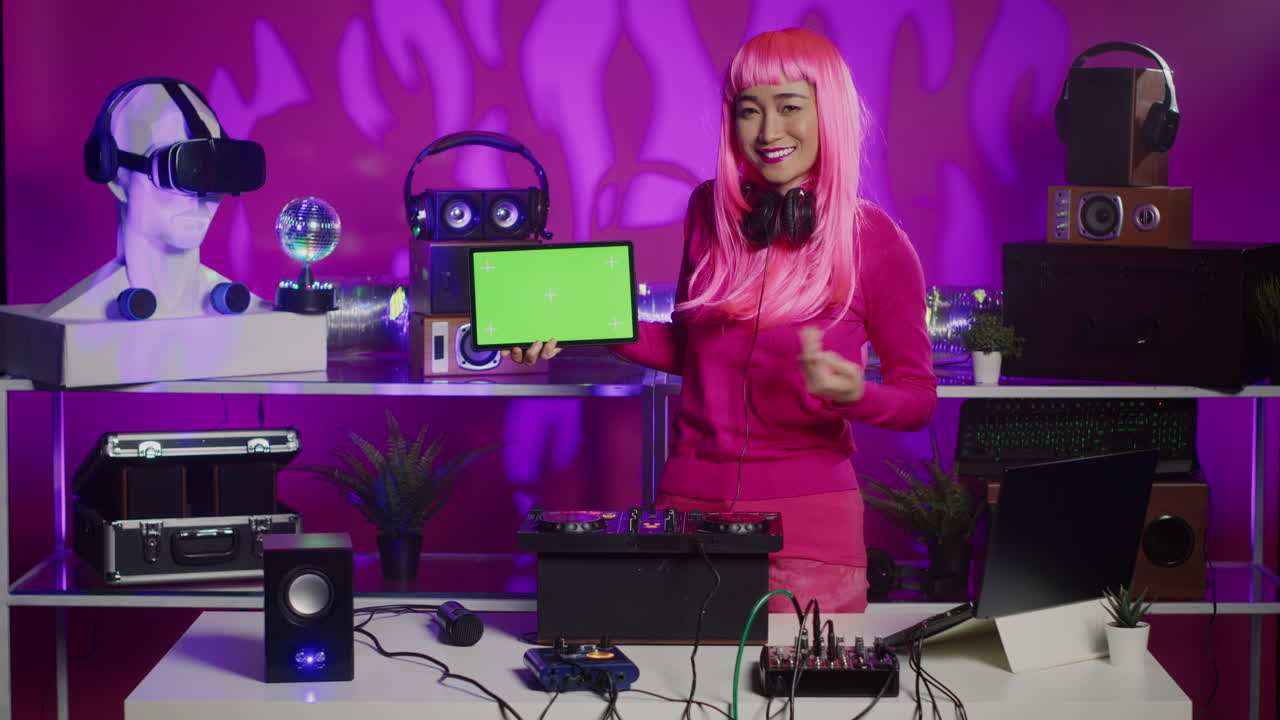 Woman with pink hair presents a tablet with green screen in a DJ setup