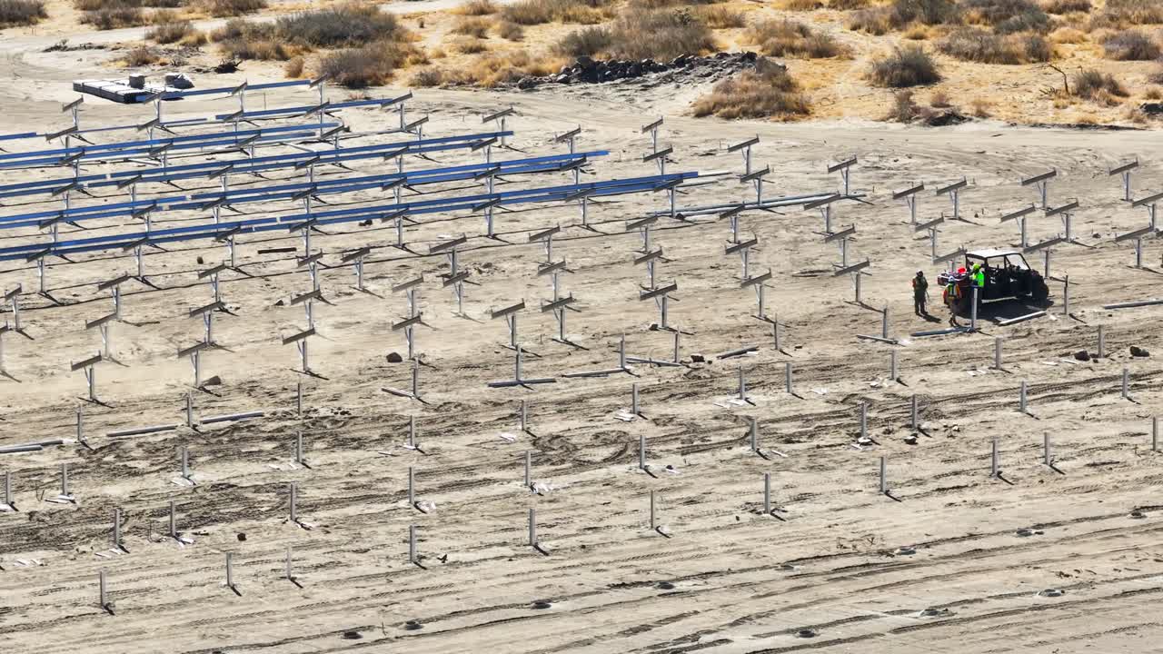 일들이 atv를 운전하면서 사막 태양 전지 건설 현장을 지나가고, 밝은 빛 하늘 아래 금속 프레임 구조의 줄로 둘러싸여 있습니다.