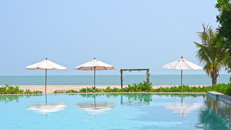 against an ocean background, beach umbrellas reflect into the water of a resort swimming pool