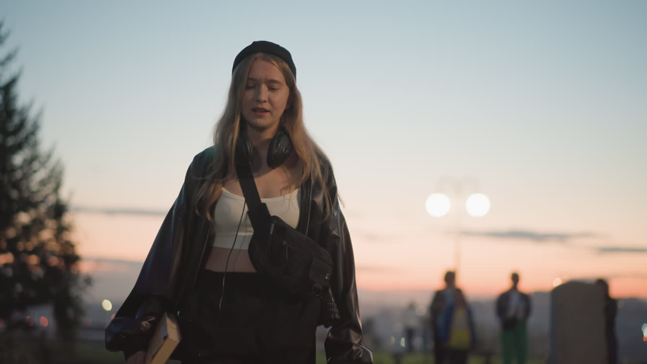 Donna che ammira il tramonto sulla città, con giacca di pelle e berretto, tiene in mano un taccuino e delle cuffie mentre si lascia avvolgere dal caldo bagliore serale e dalle luci lontane della città, in uno stato d'animo contemplativo, con un bokeh delicato