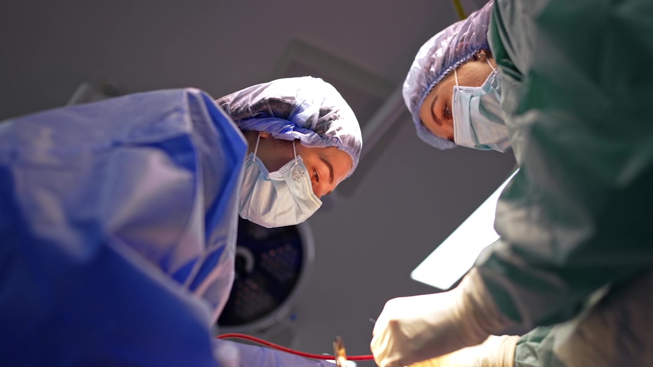 Due chirurghi chinati sul campo operatorio durante un intervento di neurochirurgia. Vista dal basso verso l'alto, camici blu e verde acqua, maschere, cuffie, lampada chirurgica e telo operatorio visibili sopra.