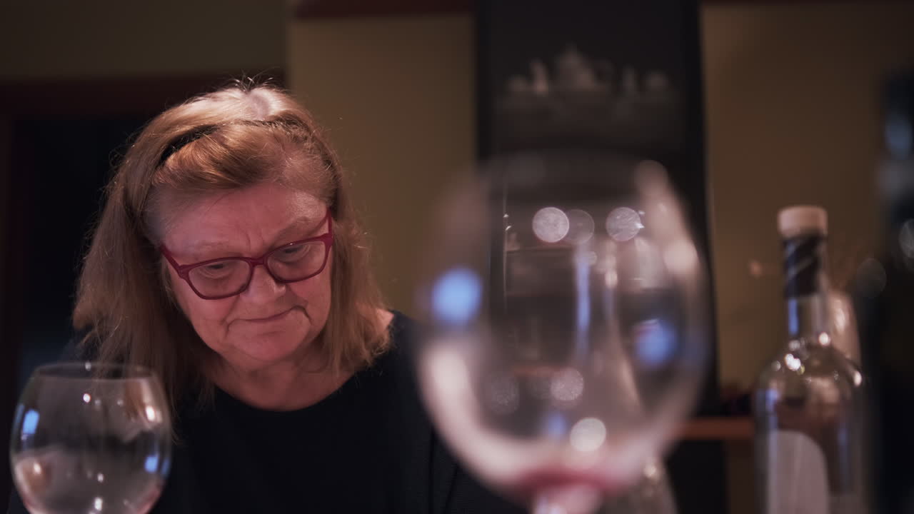 Oudere vrouw met bril zit aan de eettafel en kijkt lachend in de camera. Een gelukkige, bedachtzame senior vrouw tijdens een familiediner.