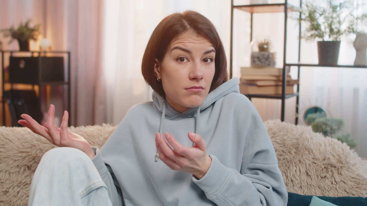 Confused caucasian woman on sofa raising eyebrows feeling embarrassed about ambiguous question