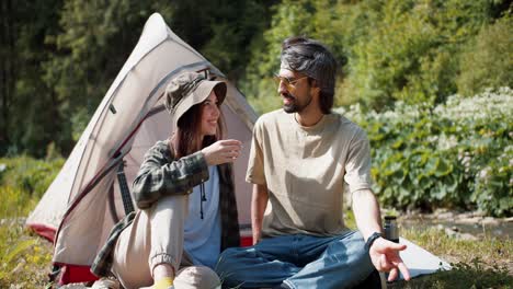 Guy-and-girl-tourists-sit-talking-near-the-tent-girl-drinks-tea-from-a-thermos.-Settlements-near-the-tent-on-the-background-of-the-forest