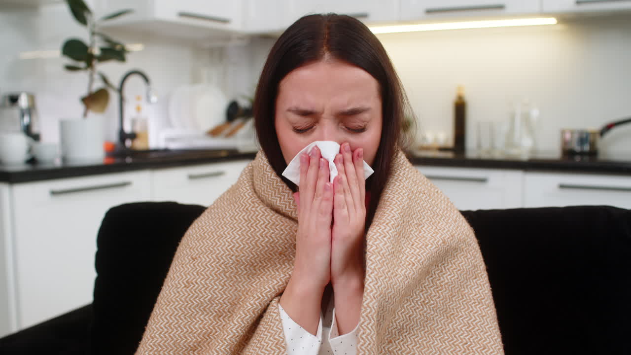 Young woman wrapped in blanket sneezes uses napkin cold flu symptoms sniffles unwell tired weak rest