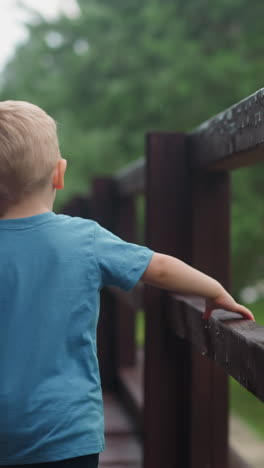 little blond child walks touching wet wooden railing on veranda deck at cottage house after warm spring rain backside view slow motion