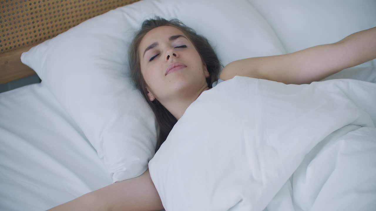 hermosa morena despertando lentamente por la mañana se estira y se levanta de la cama el sol brilla sobre ella desde la gran ventana. feliz joven saluda el nuevo día