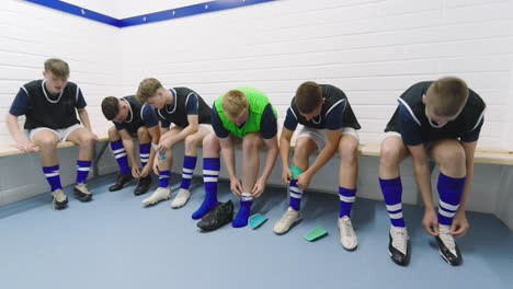 male soccer players wearing black vests and tying shoes sitting on bench in changing room