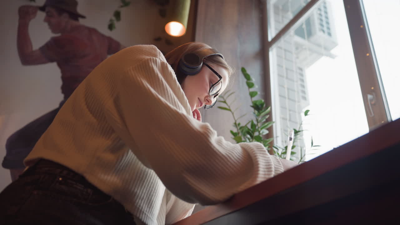 upward angle view of young woman wearing glasses and headphones while studying by window in warm indoor space with leafy plant and mural on wall creating peaceful focused atmosphere