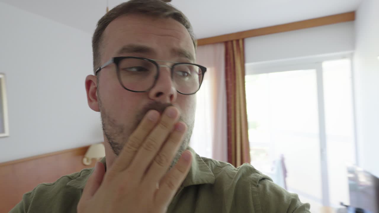 Primo piano di un uomo che si filma in una stanza d'albergo, coprendosi la bocca con una mano. La sua espressione facciale mostra paura, shock o improvvisa sorpresa.