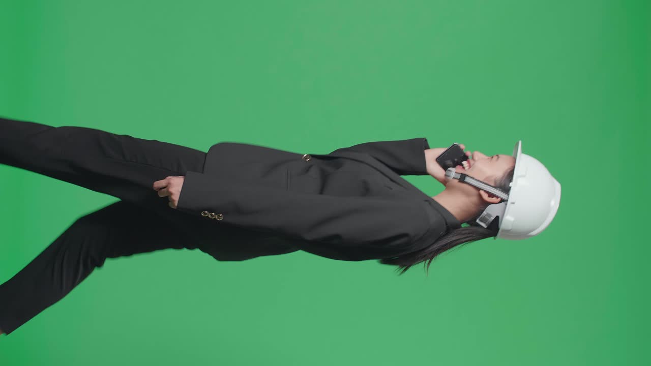Side View Of Smiling Asian Female Engineer Talking On Smartphone And Celebrating While Walking In The Green Screen Studio