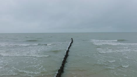 vista aérea de un antiguo muelle de madera en la costa del mar báltico, día de invierno nublado, playa de arena blanca, postes de madera, olas golpeando la costa, amplio tiro de drones avanzando bajo