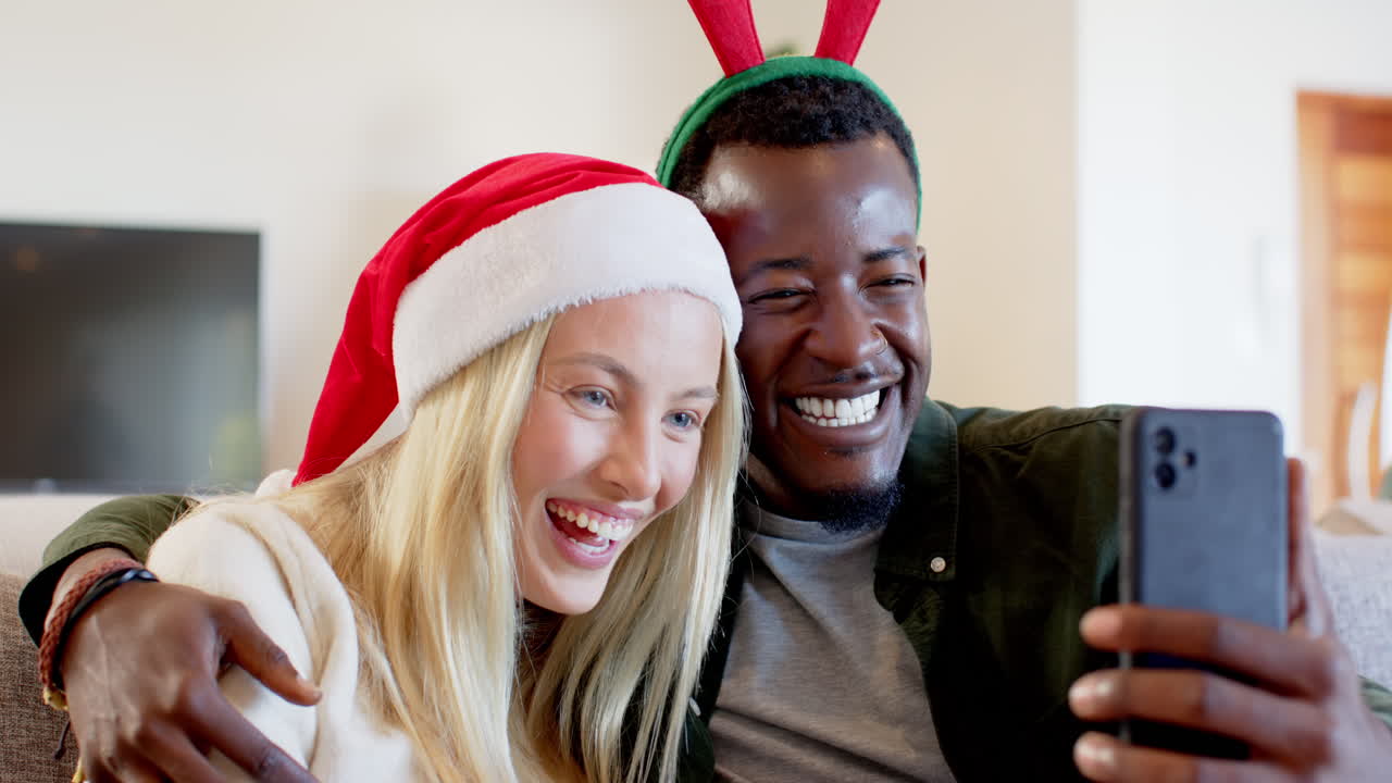 Happy diverse couple wearing festive hats taking selfie at home, celebrating Christmas joy