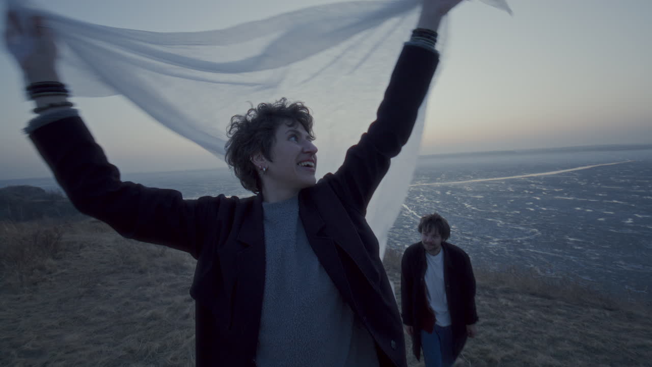 Smiling Woman Holding Fabric Overhead as Boyfriend Hugging Her by Frozen Lake