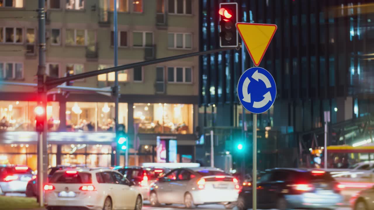 bokeh atasco de tráfico de automóviles borroso en la ciudad ocupada señal de carretera rotonda de conducción cruce de carretera por la noche. timelapse hora pico carretera del centro de la ciudad. coches irreconocibles en movimiento rápido por la noite. semáforos senderos