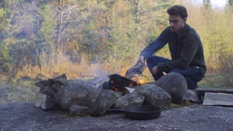 a man tends a campfire in the morning forest, placing wood on the fire
