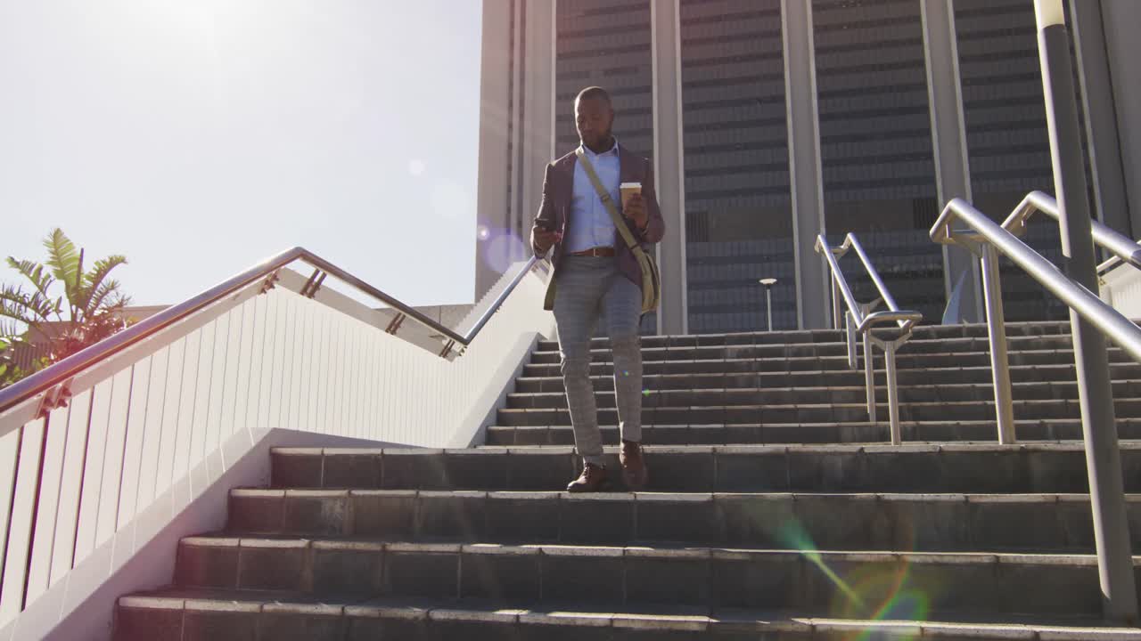 hombre afroamericano en la ciudad caminando por las escaleras, sosteniendo café, usando un teléfono inteligente