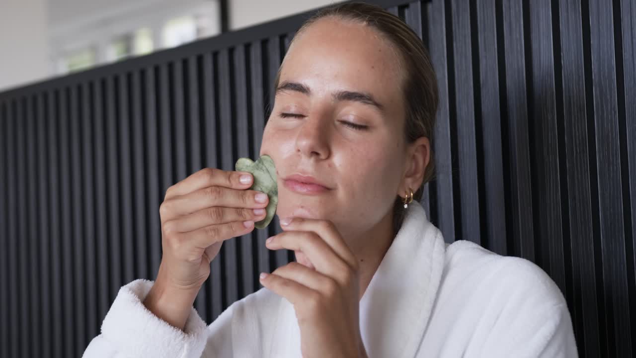 Woman wearing bathrobe placing green gua sha near jaw and massaging cheeks in bedroom for skincare