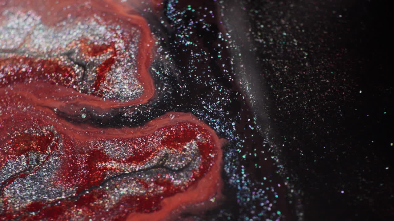 Macro shot of red, black, and grey glitter inks, as a dense black glitter stain expands and flows across the entire frame