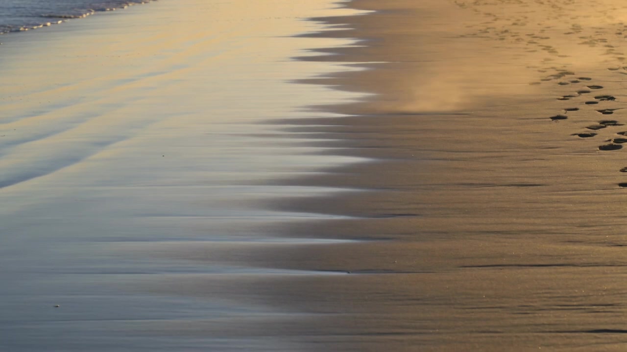 Onde che avanzano modellando increspature centrali di sabbia bagnata sulla battigia, con impronte e riflessi del sole.