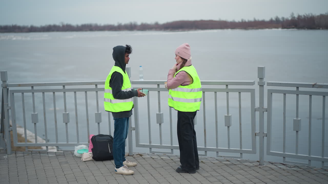 Two people wearing reflective safety vests standing on paved waterfront promenade, conversing near metal railing with river and bare trees in background, backpacks and food containers placed on ground