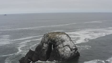 Stormy-day-aerial-of-a-rocky-ocean-shore,-panning-around-a-large-rock-in-the-ocean,-waves-crash-against-dark-sea-rocks,-the-sky-looks-dark-with-clouds