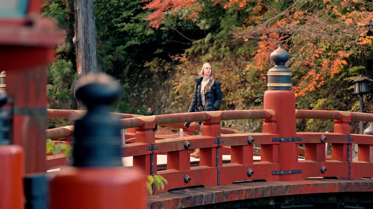 A cinematic video capturing a woman gracefully walking across the historic Shinkyo Bridge in Nikko, Japan, surrounded by the warm hues of autumn foliage.