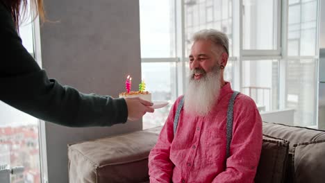 Un-Hombre-Feliz-De-Cabello-Gris-De-Edad-Avanzada-Con-Una-Barba-Exuberante-Con-Una-Camisa-Rosa-Recibe-Un-Pastel-Y-Velas-De-Su-Hija-Adulta-Con-Una-Chaqueta-Verde-En-Un-Apartamento-Moderno.