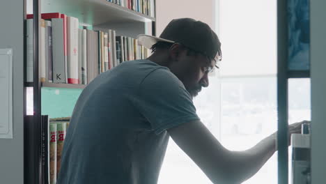 close up side view of young man wearing cap in library focused on sorting through books on shelf, with natural daylight streaming from large window