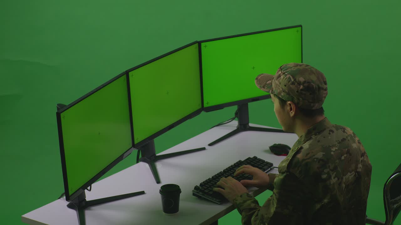 vista lateral de un soldado asiático trabajando con un simulacro de varios monitores de computadora en un estudio de pantalla verde