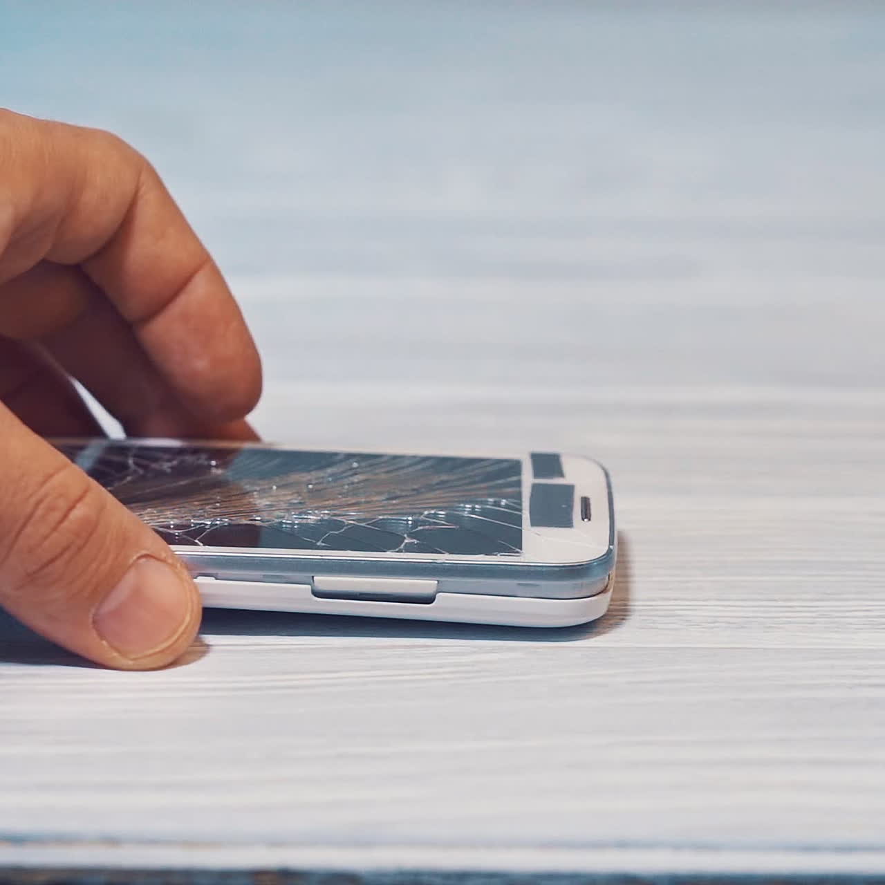 Male's hand touching the cracked white smartphone on the grey wooden surface. Broken mobile phone in the hand of a man. Close-up