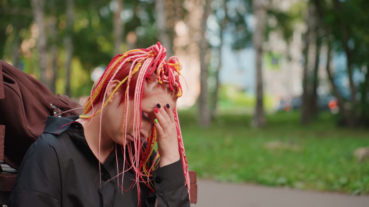 Mujer blanca con rastas rosas, cubriendo su cara con la mano y luego bajando la cabeza, sentada en un banco del parque, tráfico distante difuminado detrás de los árboles, secuencia íntima de dolor privado y respiración lenta