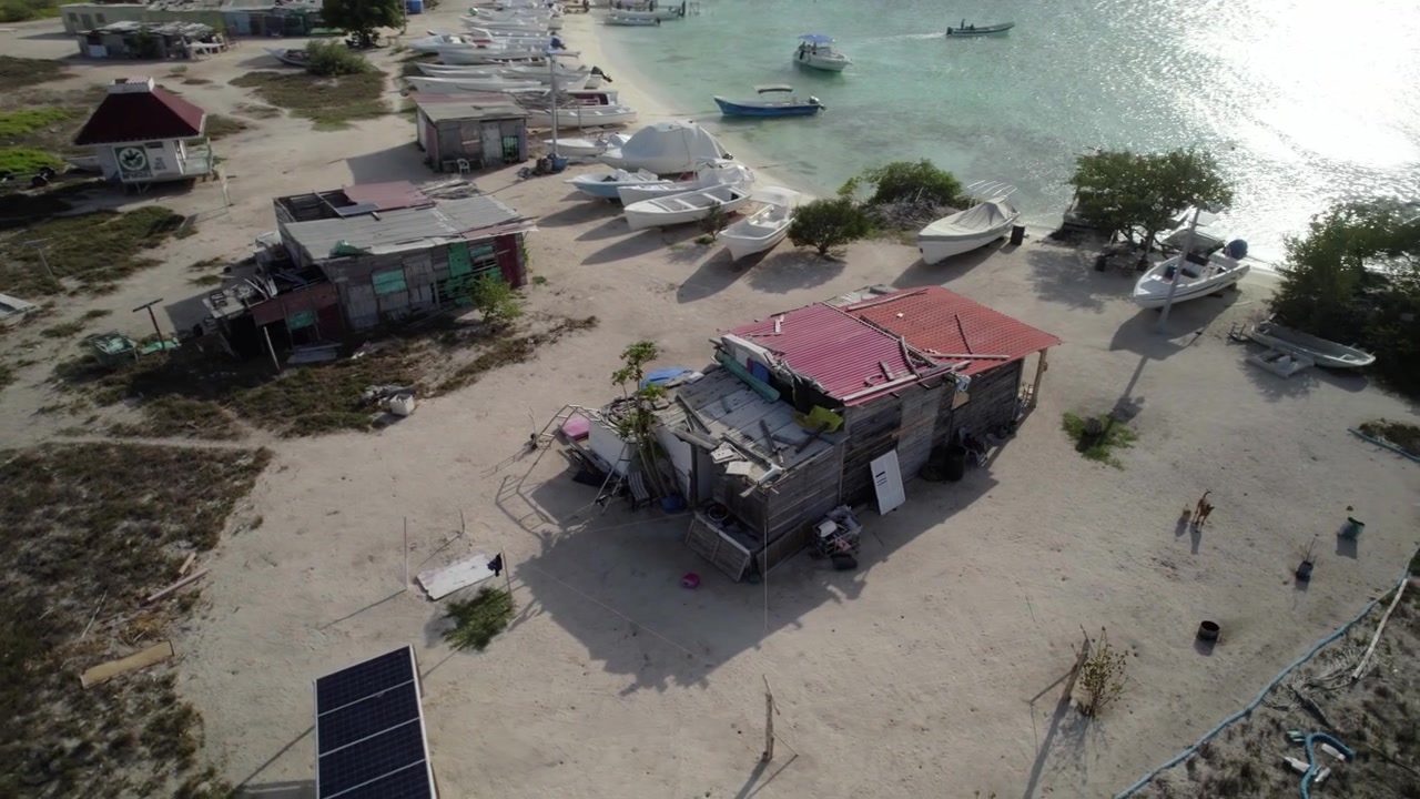 Un'ampia ripresa drone rivela un umile rifugio in legno con tetto di tegole rosse, circondato da barche tranquille e acque turchesi limpide in un ambiente costiero remoto. Los Roques Venezuela 4K DLOG