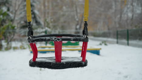 el columpio de los niños congelados cubierto de hielo y hielo colgando a lo largo de su marco rojo y negro, suspendido por cuerdas amarillas, columpio suavemente establecido contra un fondo de patio de recreo nevado