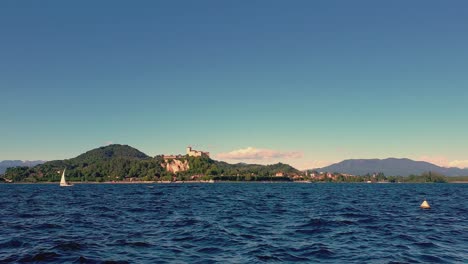 pequeño velero navegando en el lago maggiore aguas agitadas con montañas y castillo de angrya en el fondo, italia