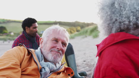 Loving-Senior-Couple-Relaxing-By-Fire-With-Adult-Offspring-On-Winter-Beach-Vacation