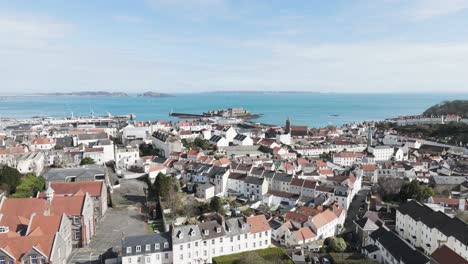 High-level-flight-over-St-Peter-Port-roof-tops-out-to-sea-over-harbour-and-Castle-Cornet-with-views-of-islands-on-sunny-day