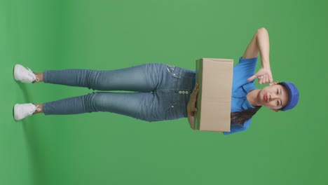 full body of unsatisfied asian female courier in blue uniform showing thumbs down gesture while delivering a carton on green screen background in the studio